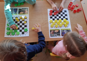 Na stole leżą plansze do gry w warcaby. Na nich leżą klocki w kolorach czerwonym, żółtym i zielonym z zakrętek do butelek. Dzieci grają w warcaby.